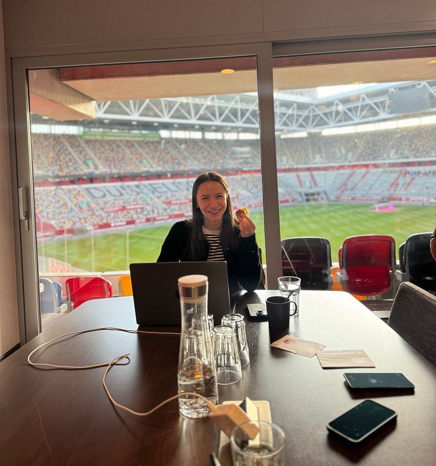 Paula sitzt mit Laptop in einer Stadion-Loge, im Hintergrund das Spielfeld. Sie arbeitet fokussiert und lächelt in die Kamera.